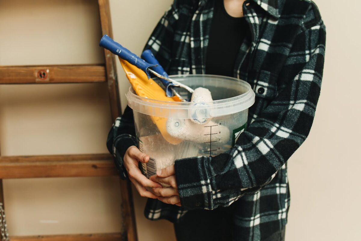 Close-up of a person holding a bucket filled with renovation tools, including paint rollers and scrapers.
